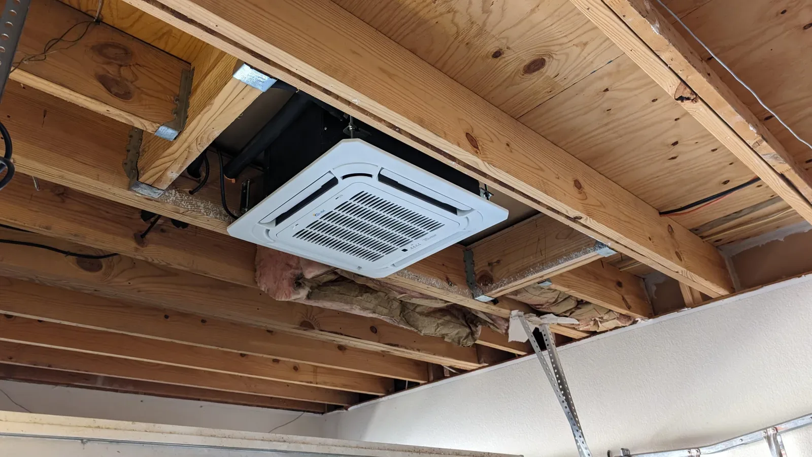 White ceiling cassette mini split head recessed between exposed wood floor joists, the kind of in-ceiling install a DIY cassette project involves