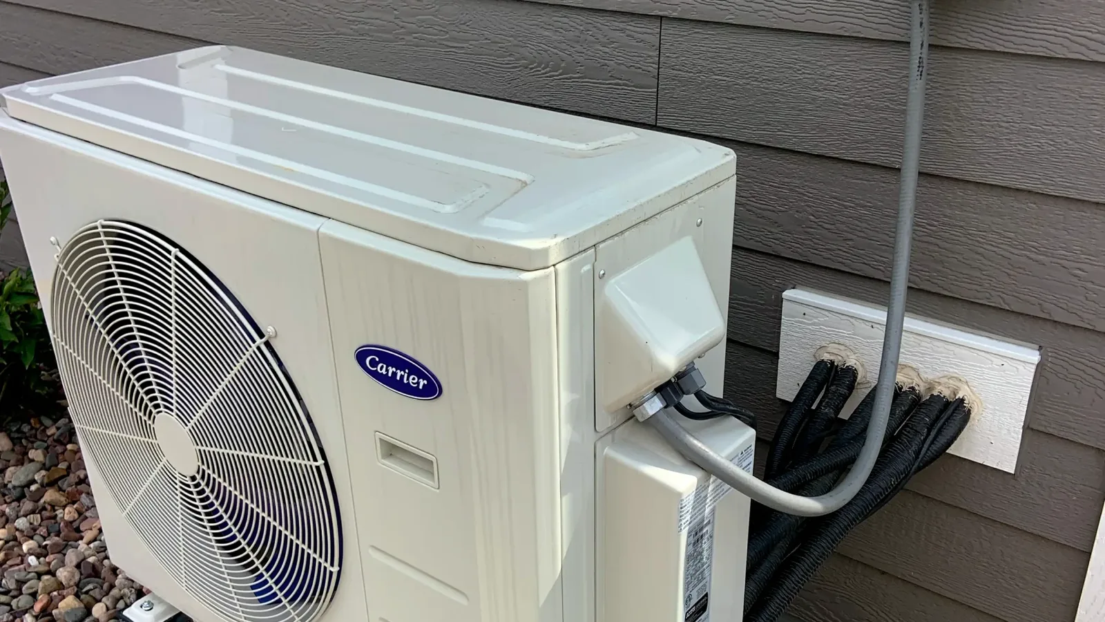 Multi zone mini split outdoor unit on a stand beside a house, with three insulated line sets running from the wall plate into the condenser and a disconnect box above