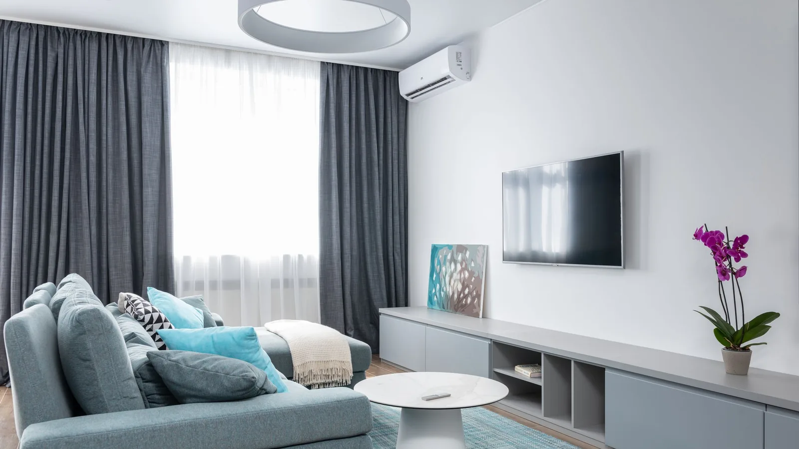 White wall-mounted mini split heat pump indoor unit above the curtains in a living room with a blue sofa, the kind of single-room DIY mini split install this guide covers