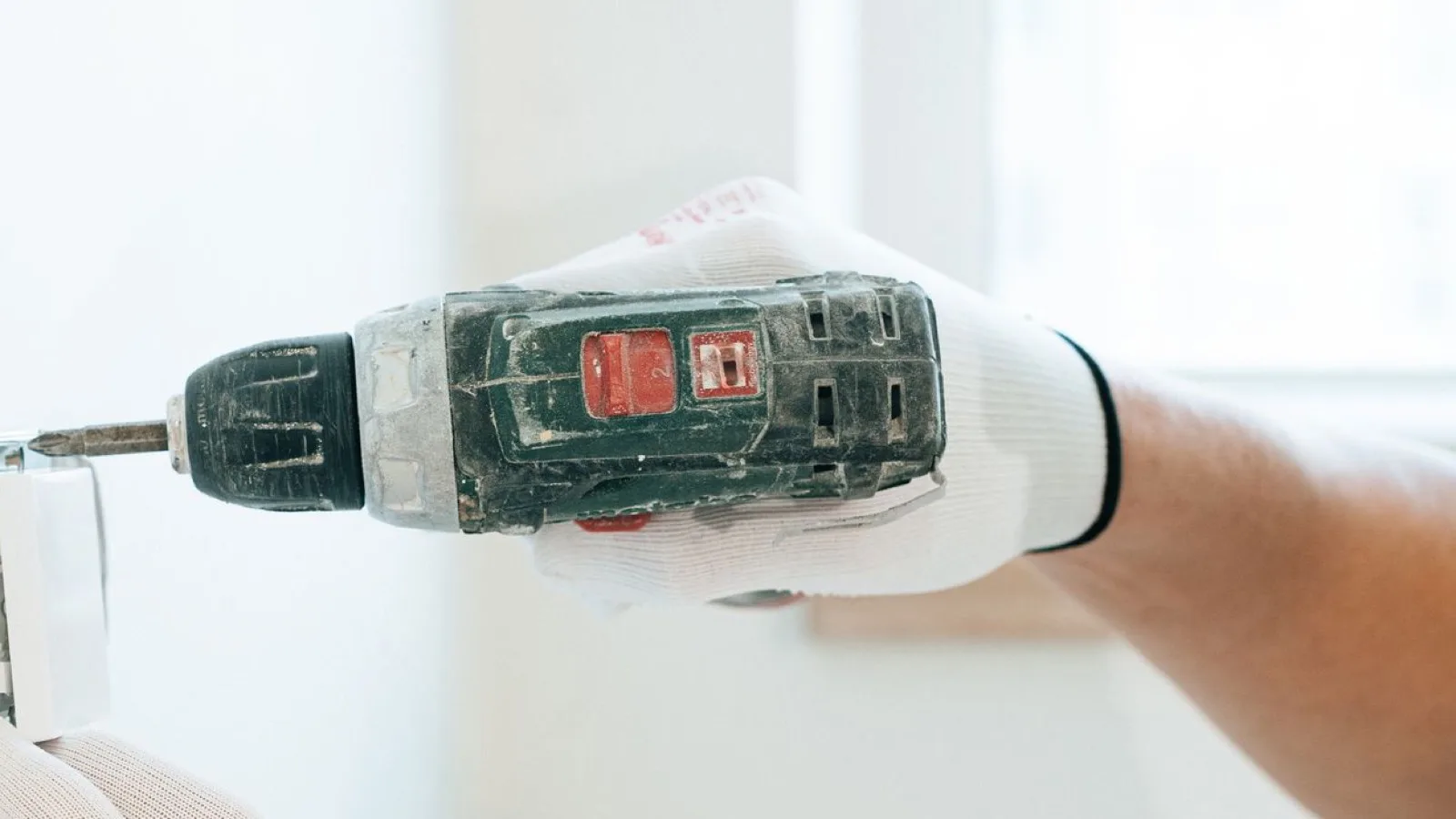 Gloved hand driving a screw into a white interior wall with a cordless drill, the same step as fixing a mini split mounting plate
