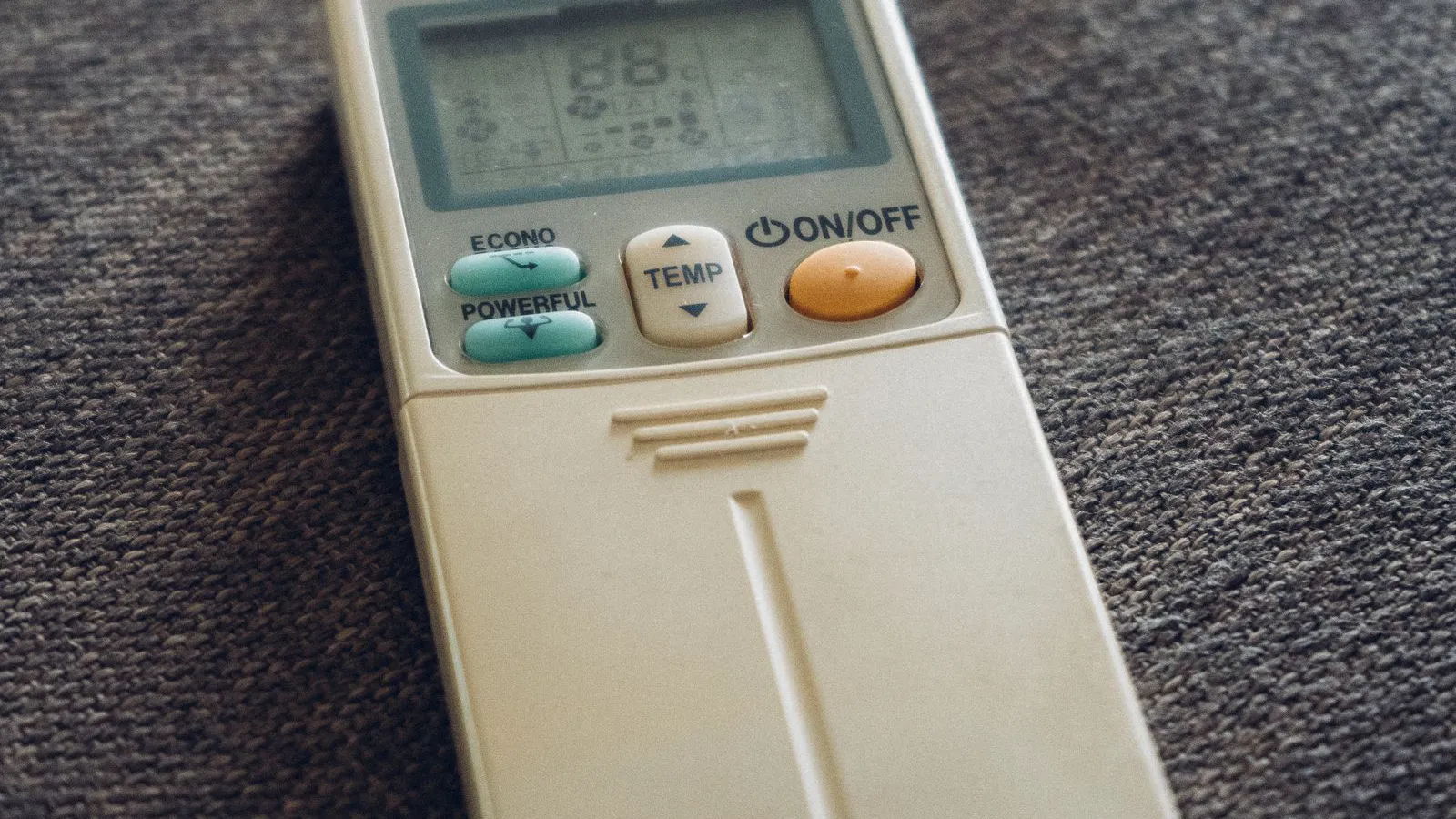 Beige ductless air conditioner remote with Econo, Powerful, Temp and On/Off buttons lying on a gray couch, used to switch to dry mode on a mini split
