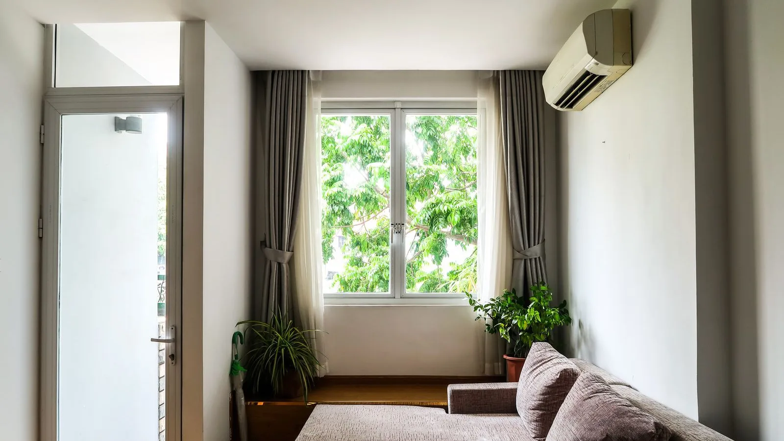 Bright living room with a sofa, curtained window and houseplants, and a wall-mounted ductless mini split head high on the right-hand wall