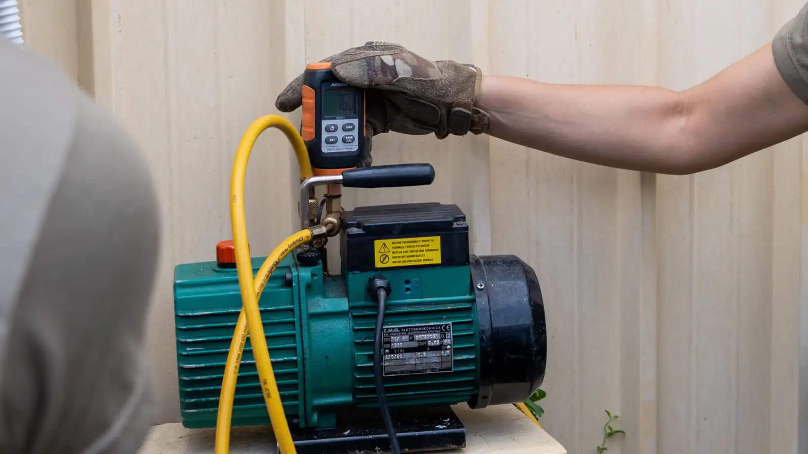 Gloved hand holding a digital micron gauge on a green vacuum pump with a yellow hose, the vacuum step that a no-vacuum DIY mini split with a pre-charged line set skips