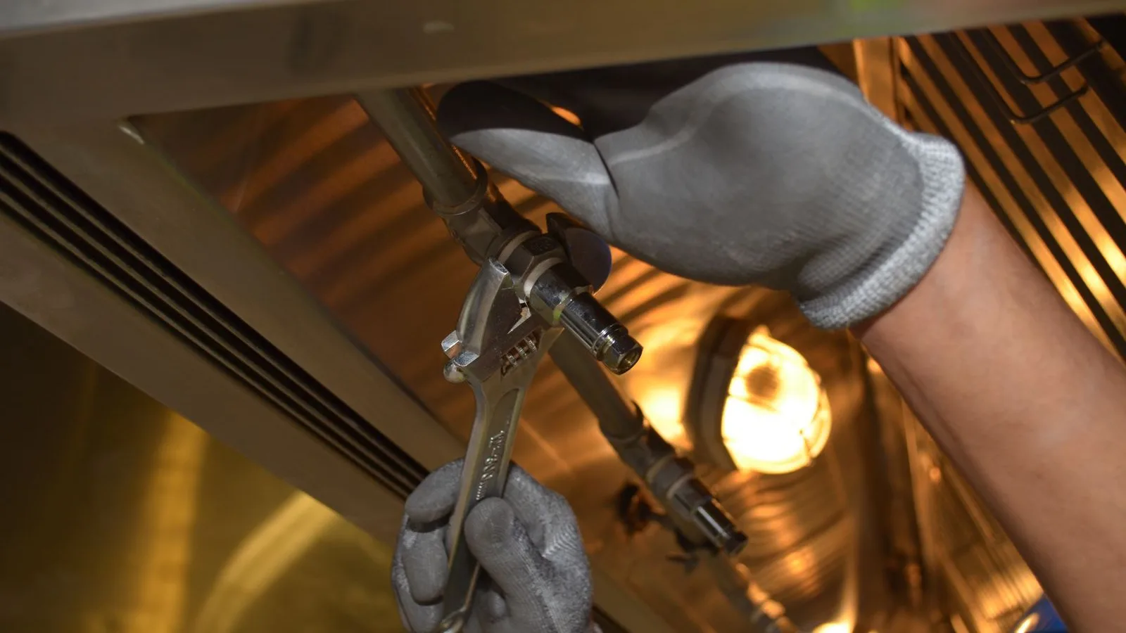 Gloved hands holding an adjustable wrench on a threaded refrigerant fitting, the two-handed grip used when tightening a quick connect line set coupling