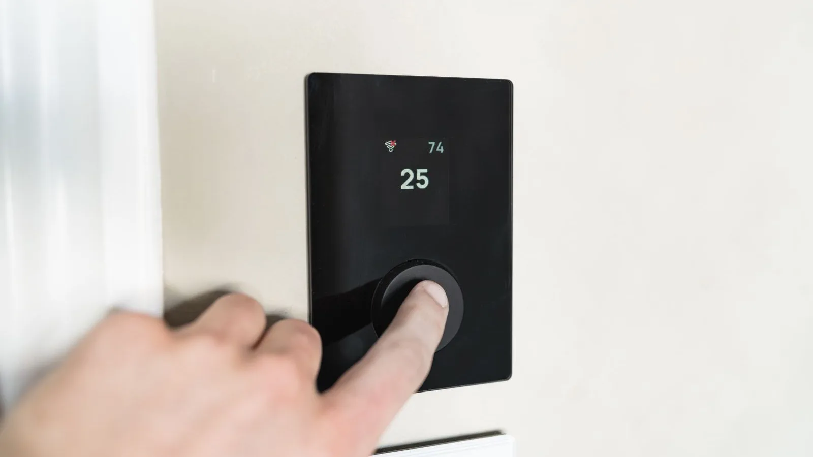 A finger pressing the round dial on a black glass wall thermostat showing a set temperature